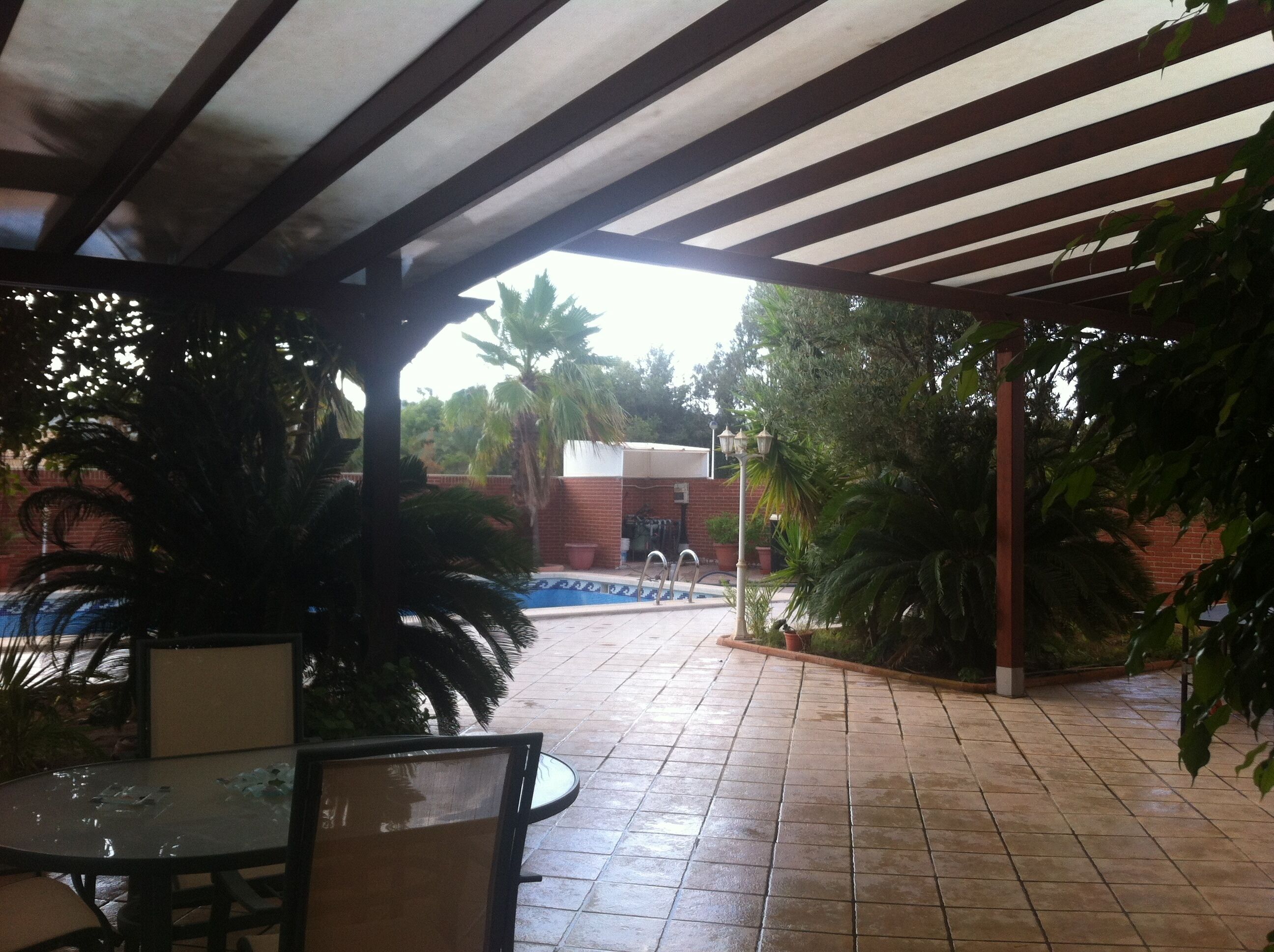 A covered patio area with a tiled floor, a pool, and surrounding greenery.