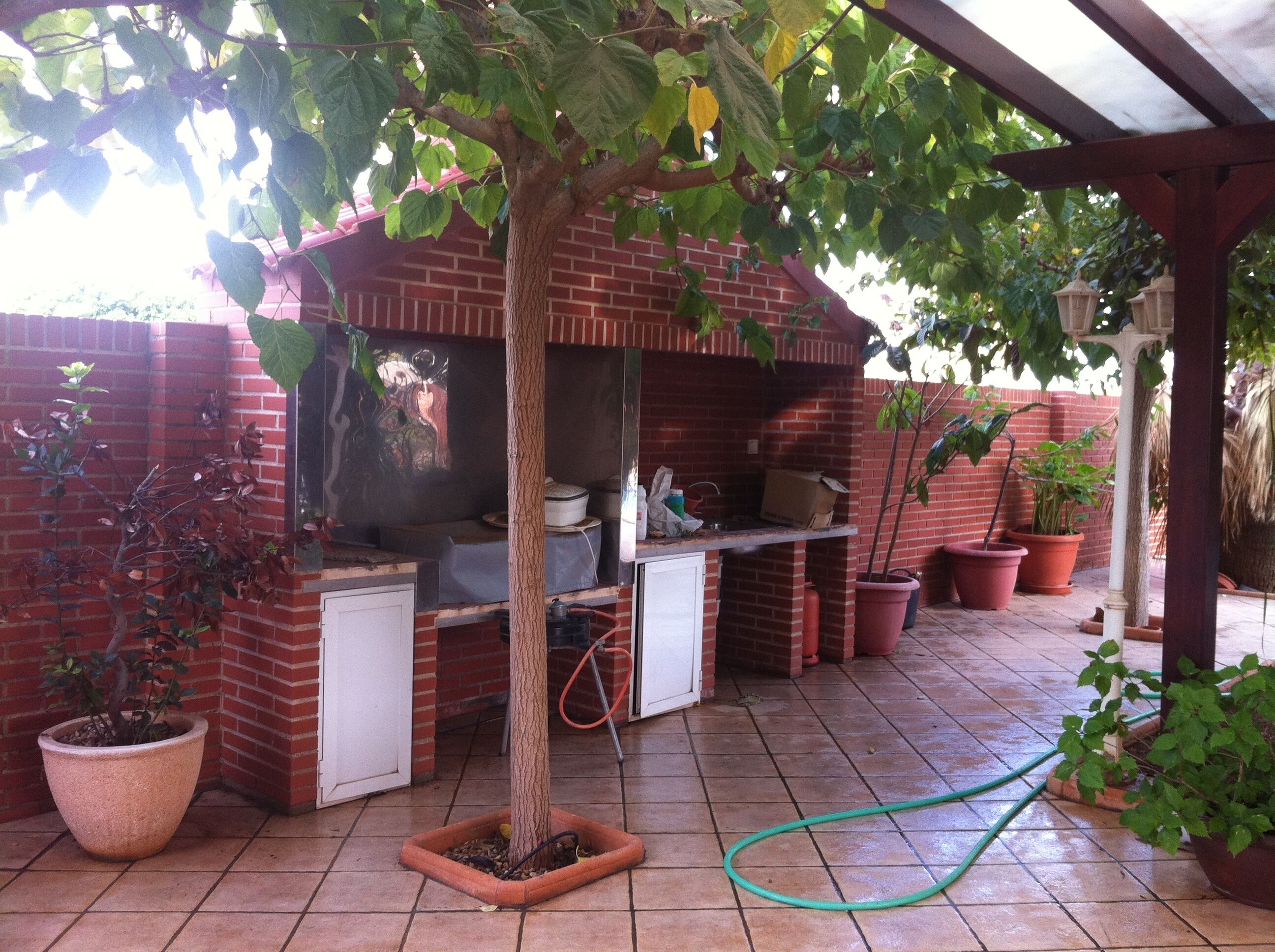 A brick barbecue area with a tiled patio, potted plants, and a hose.