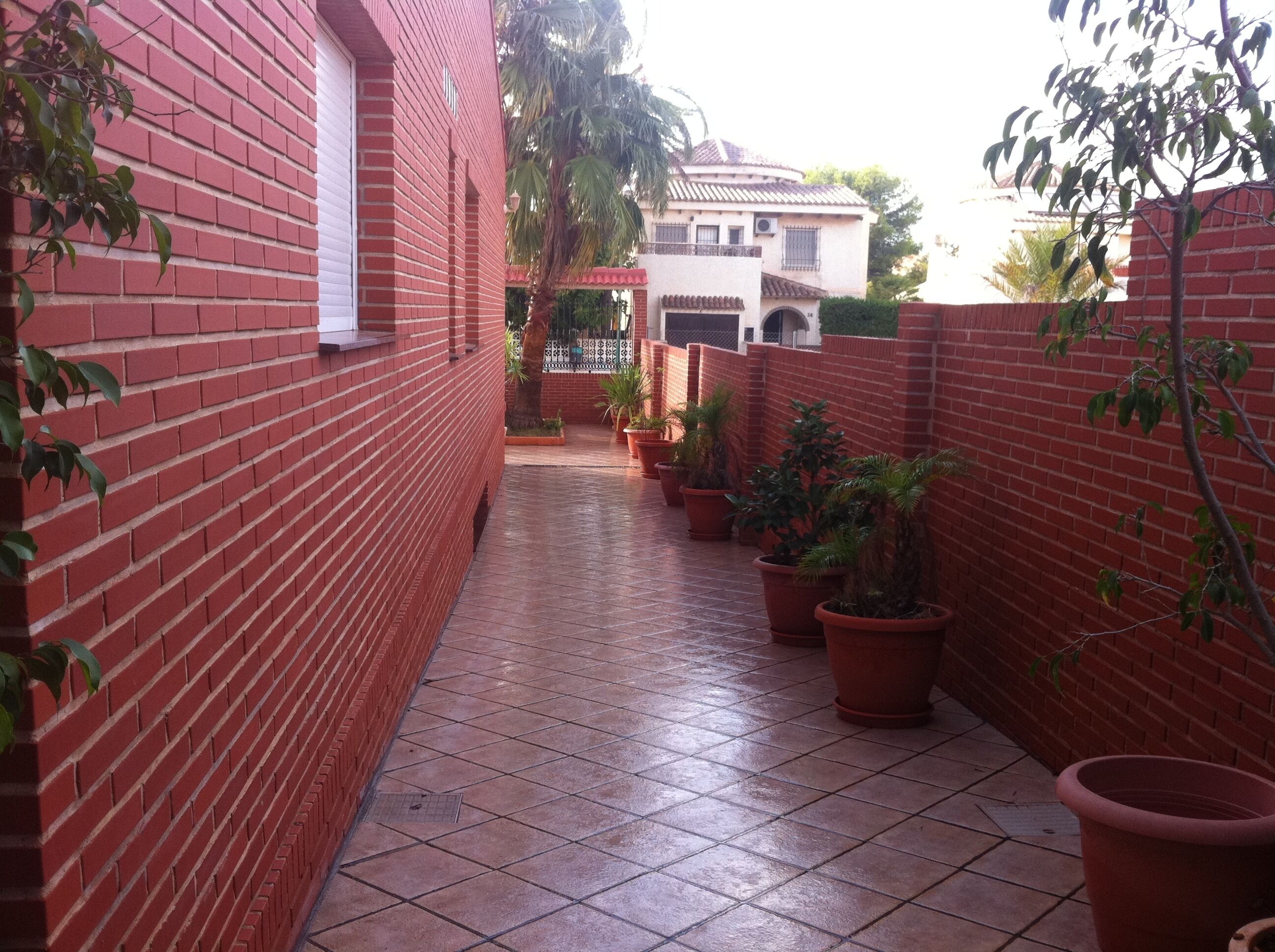 A narrow brick corridor with tiled flooring, potted plants, and a building in the background.