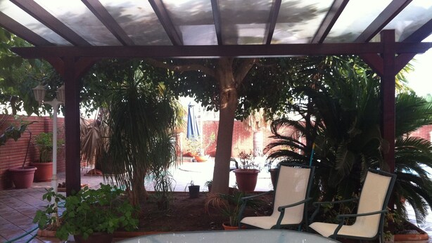 Un patio couvert avec une table en verre et deux chaises, entouré de plantes en pot et bordé d’un mur en briques au fond.