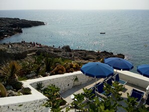 Une vue sur la côte, avec un paysage rocheux, des cactus et une plage agrémentée de parasols et de chaises bleues.