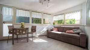 Una habitación con un sofá marrón, dos sillones, una mesita con un jarrón y ventanas grandes con cortinas.