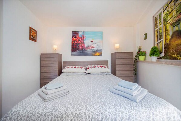 A neatly made bed with a patterned bedspread and pillows, two bedside tables with lamps, a wall-mounted painting, and a window with a view of greenery.