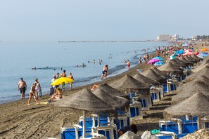 Uma praia com guarda-sóis de palha e espreguiçadeiras azuis, areal arenoso e mar azul cristalino.