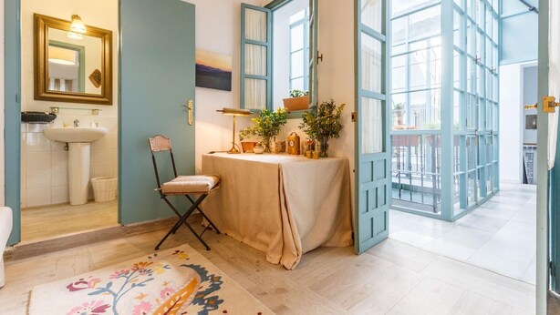 A bathroom with a sink, mirror, and chair. There is a table with plants and a lamp. The room has a large window and a door leading to another room. - IN OFFER - LUXURY HOUSE WITH TERRACE IN LA JUDERIA, PAX 10 (SEVILLA)
