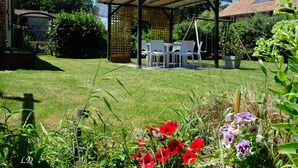 A covered outdoor seating area with white chairs and a table, surrounded by lush greenery and flowering plants. - Gîte with Jacuzzi in the countryside 18 kms from the ocean (La Boissière-Des-Landes)