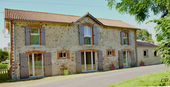 A two-story stone house with a tiled roof, multiple windows with shutters, and a paved driveway.
