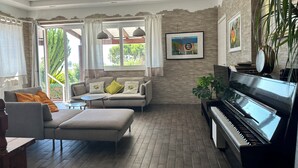 Un salon comprenant un canapé, un pouf, un piano et donnant sur un espace verdoyant.