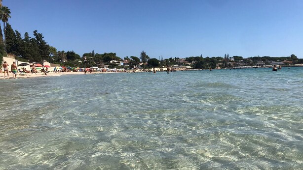 Une plage avec une eau claire et peu profonde, des gens qui prennent des bains de soleil et des bâtiments en arrière-plan.