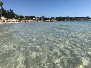 Une plage avec une eau claire et peu profonde, des gens qui prennent des bains de soleil et des bâtiments en arrière-plan.