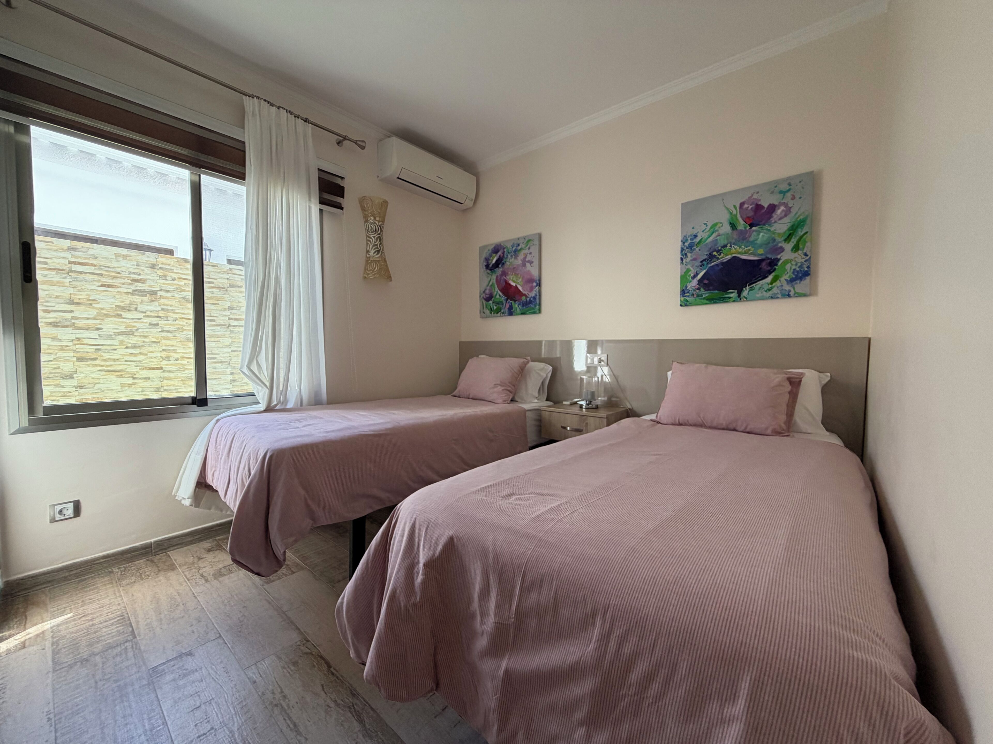 A hotel room with two beds, a window with curtains, an air conditioning unit, and two floral paintings on the wall.