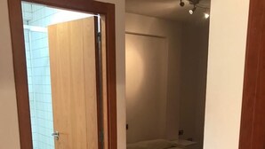 Una habitación con piso de madera, una cama, una mesita y una puerta que da a un baño con azulejos blancos.