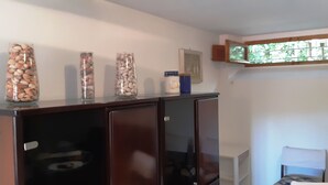 A room with a wooden cabinet topped with three glass jars containing nuts, a window with a view of greenery, and a small white shelf with a blue container. - Apartment with garden in Castiglioncello (Rosignano Marittimo)