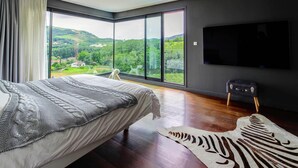 A modern bedroom with a large bed, a flat-screen TV, and a view of a green landscape through floor-to-ceiling windows.
