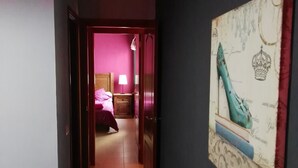 Un pasillo que conduce a un dormitorio con pared rosa y cama con manta rosa. Hay un cuadro en la pared y una puerta a la izquierda.