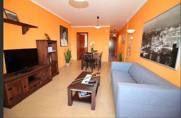 Una sala de estar con televisor, mueble de madera para entretenimiento, un sofá, una mesa de centro y un comedor con sillas y una mesa.