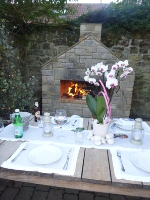 An outdoor dining setup with a stone fireplace, white orchid flowers, and a table set for two with plates, cutlery, and glasses. - Fossil Bank, a luxury detached retreat in the heart of Alnmouth, with parking (Alnmouth)