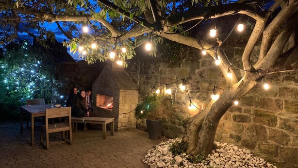 An outdoor patio with a stone wall, a brick table, and a tree with string lights. - Fossil Bank, a luxury detached retreat in the heart of Alnmouth, with parking (Alnmouth)
