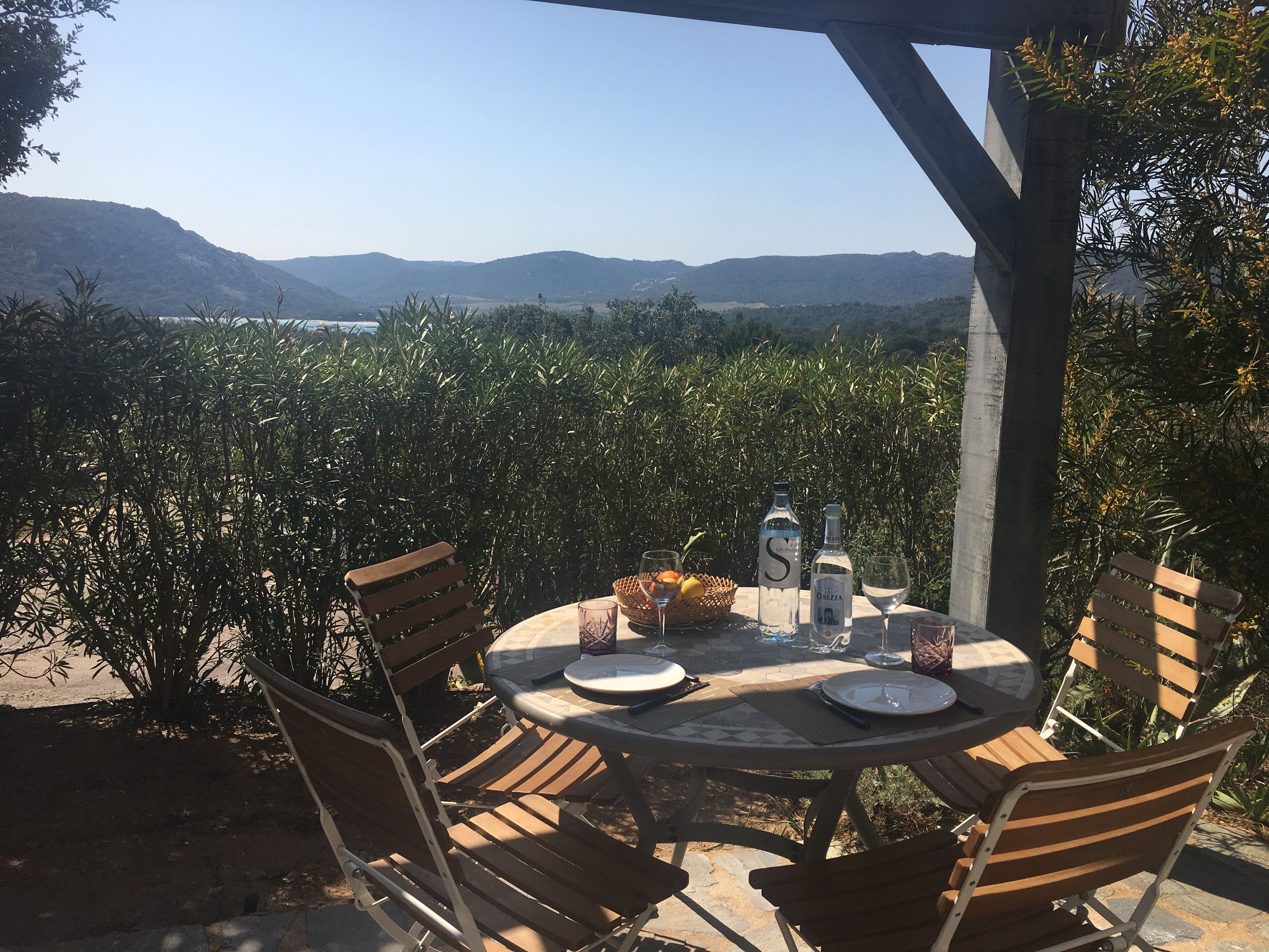 An outdoor dining area with a round table and chairs, surrounded by lush greenery and a view of distant hills.