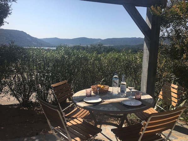 An outdoor dining area with a round table and chairs, surrounded by lush greenery and a view of distant hills. - Mini Villa with sea view terrace, 5 minutes on Spia's all inclusive rates (Porto-Vecchio)