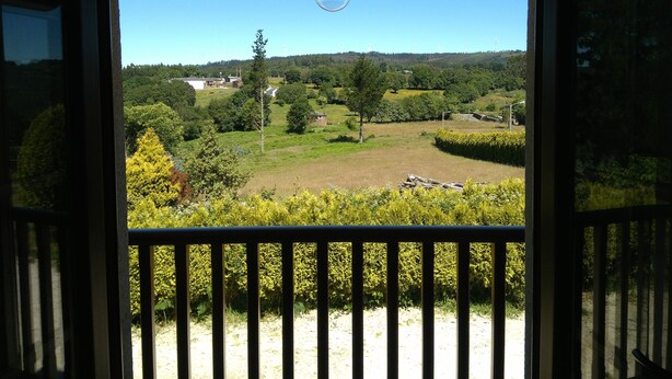 A view from a balcony with a wooden railing, overlooking a green landscape with trees and a clear sky.