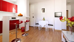 A modern kitchen with red cabinets, a microwave, and a fruit bowl. There is a dining area with a table set for two, a TV, and a vase with flowers.