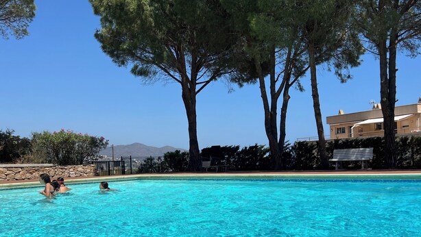 A clear blue swimming pool surrounded by trees and a stone wall, with a bench and a building in the background. - Apartment Llanca Sea view, Swimming pool and 5min walk to the beach (Llanca)