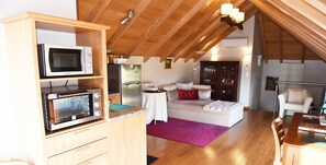 A cozy, well-lit attic room with wooden beams, a dining area, and a kitchenette.