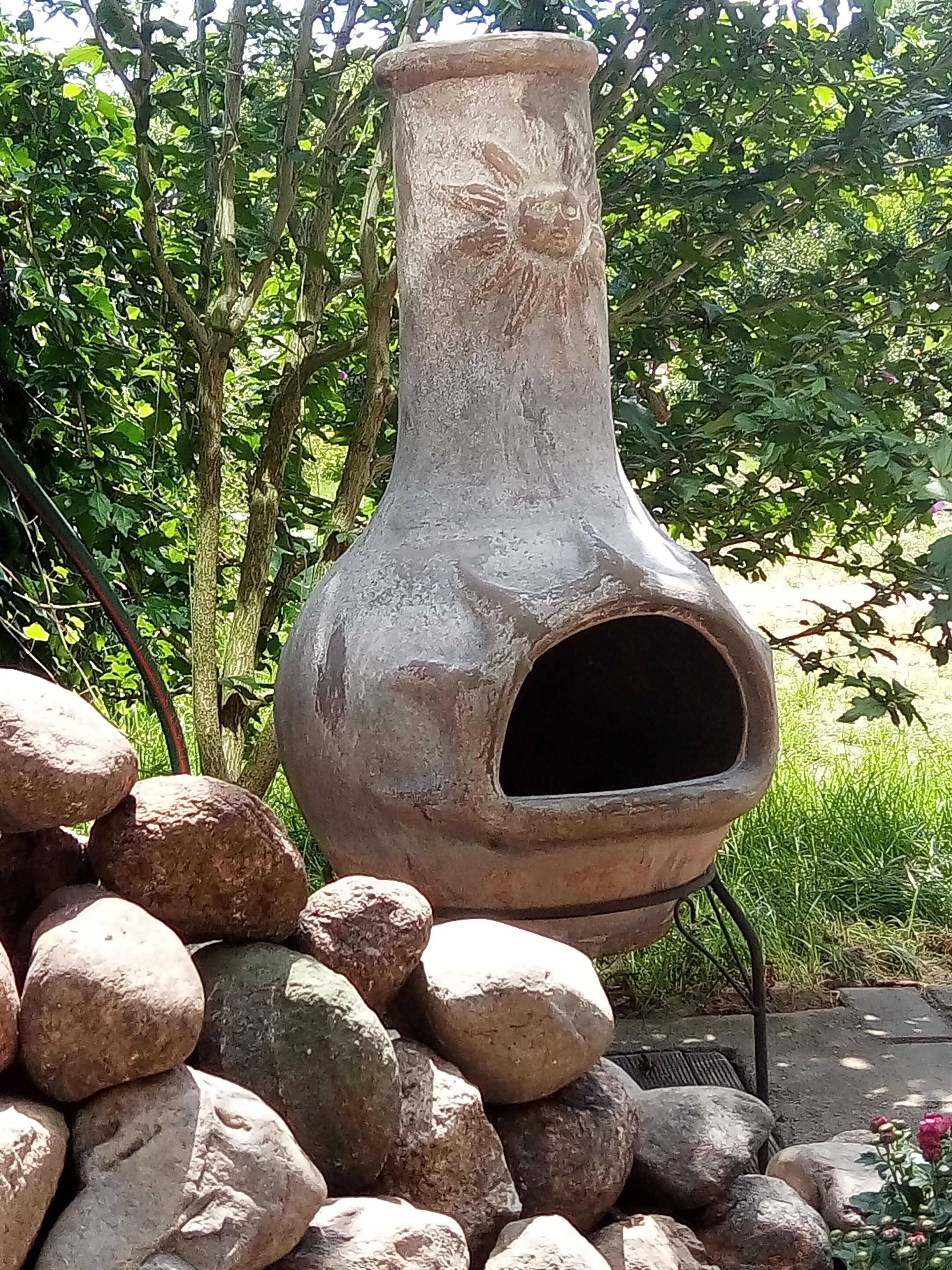 A large, rustic clay pot with a flower design, placed outdoors among rocks and greenery.