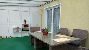 Un patio con una mesa de vidrio, sillas de mimbre, una planta en maceta y un armario blanco con estantes.