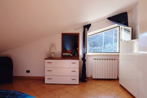Une chambre avec une commode, un miroir, un radiateur et une fenêtre donnant sur des arbres.