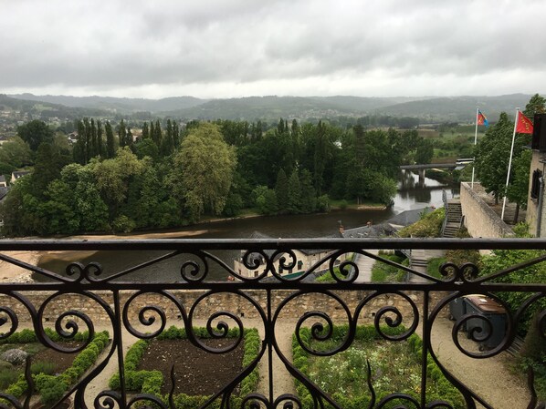 Une vue sur la rivière depuis un balcon doté d’une rambarde en métal, d’un mur en pierre et d’un pont visible au loin.