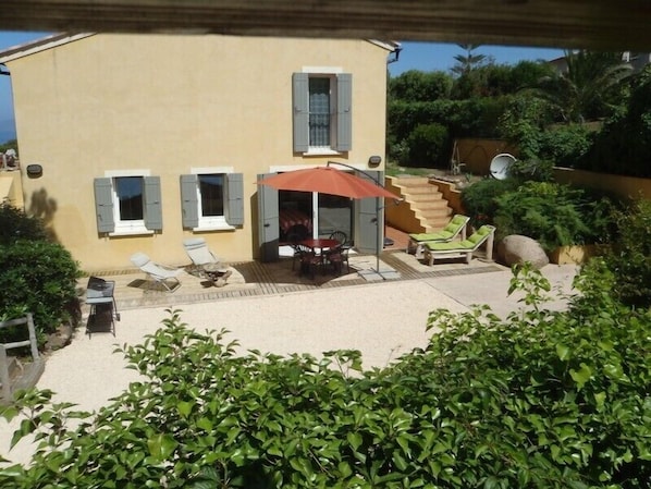 A yellow building with a patio area featuring a table, chairs, and a red umbrella. There are lounge chairs and a staircase leading to an upper level.
