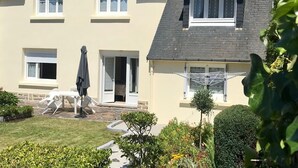 Ein zweistöckiges Haus mit geneigtem Dach, einer kleinen Veranda und einem Garten mit verschiedenen Pflanzen.