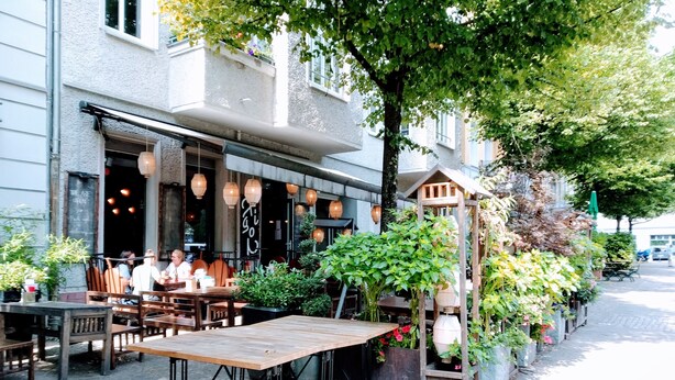 Een terras met tafels en stoelen, omgeven door potplanten en een houten pergola.