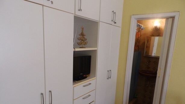 Quarto com armários brancos, uma televisão em uma prateleira e um banheiro visível através de uma porta aberta.