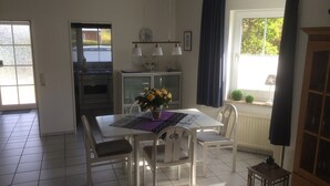 Un coin repas avec une table et des chaises blanches, un vase de fleurs jaunes et une vue sur l’extérieur par une fenêtre.