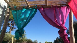 Uma pérgula de madeira com teto de tecido trançado e duas cortinas coloridas amarradas aos vigários.
