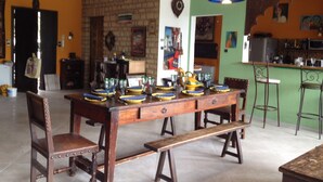 Uma sala de jantar com uma mesa de madeira posta para seis pessoas, rodeada por cadeiras. Um banco está colocado em frente à mesa. O ambiente possui uma parede de tijolos e uma parede verde, com uma área de cozinha visível ao fundo.