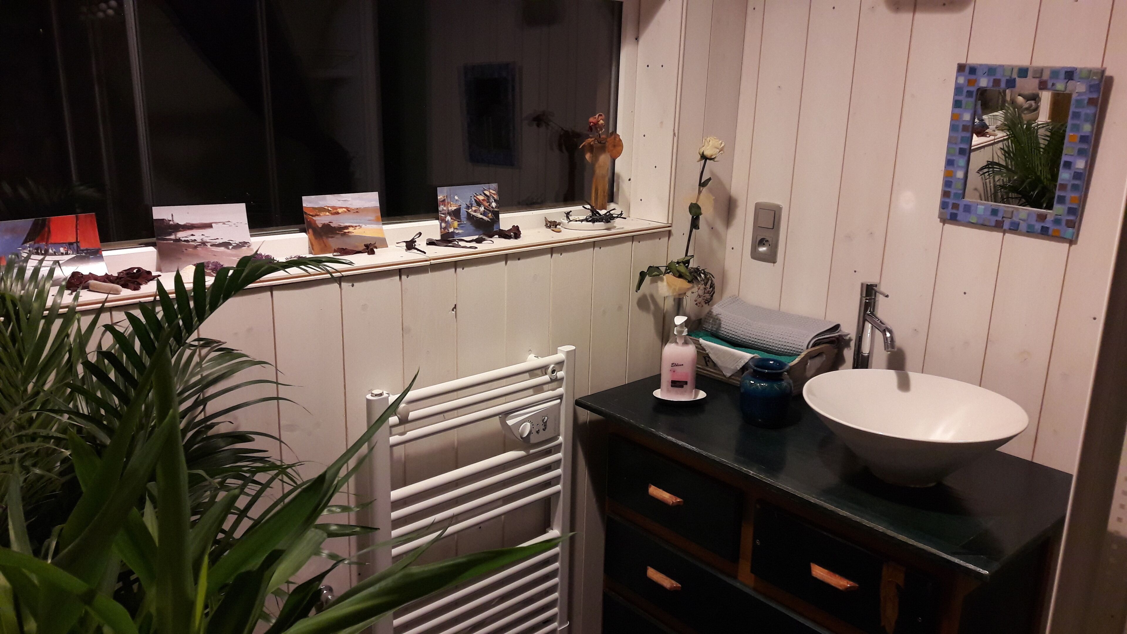 A bathroom with a white sink, a mirror with a mosaic frame, and a towel rack. There is a potted plant in the corner and a window with a view of the outside.