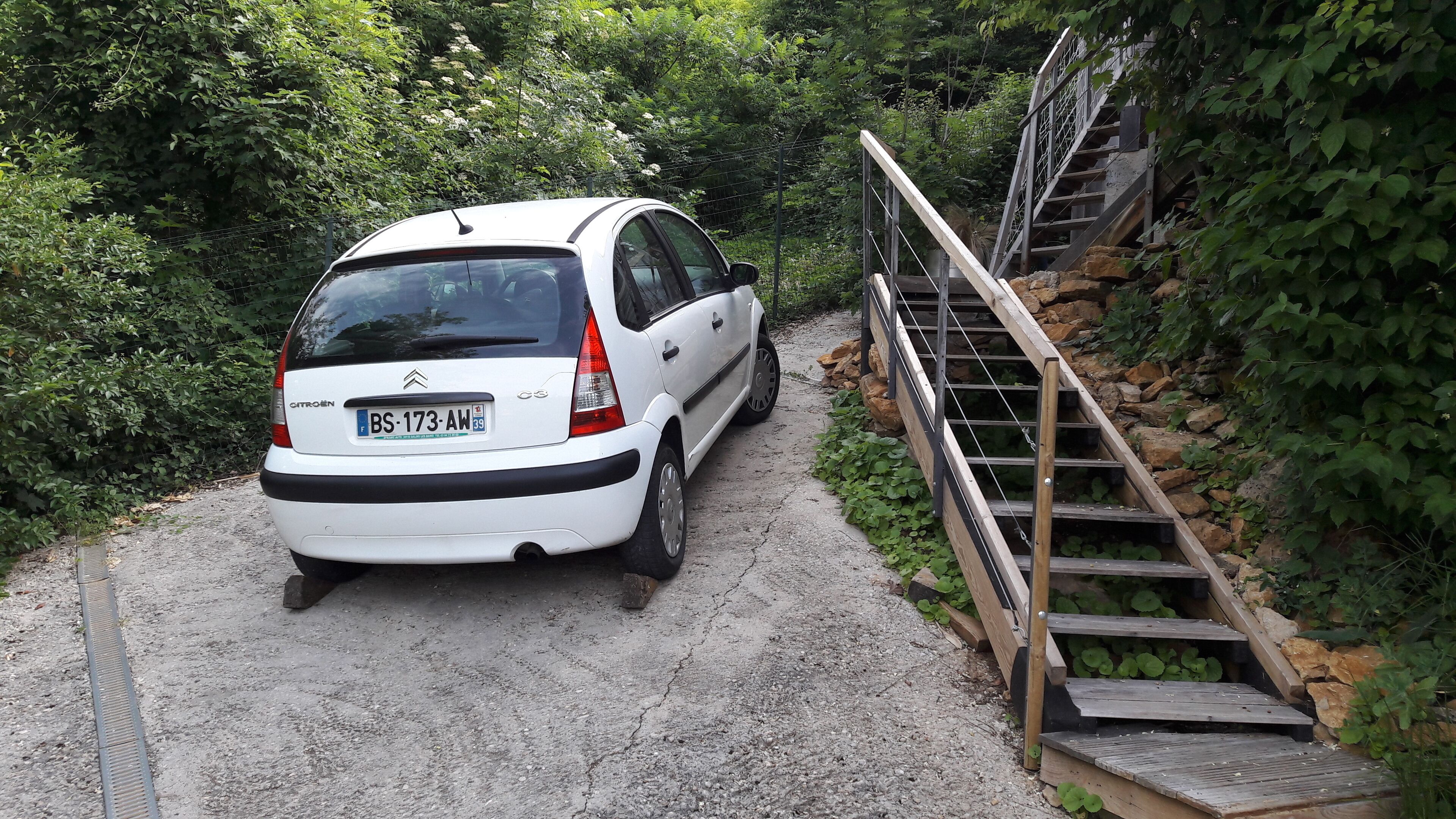 A white Citroën car parked on a gravel path beside a wooden staircase leading up a rocky hill.