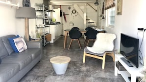 Una sala moderna con un sofá gris, un sillón blanco y mullido, y un televisor sobre un mueble blanco. Se observa un área de comedor con una mesa y sillas, y una cocina al fondo.