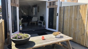Una puerta corredera abierta que da a un porche cubierto con una mesa de madera, dos plantas en macetas y un pequeño objeto naranja sobre la mesa.