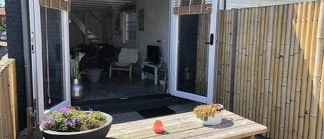 An open sliding door leading to a covered patio area with a wooden table, two potted plants, and a small orange object on the table.