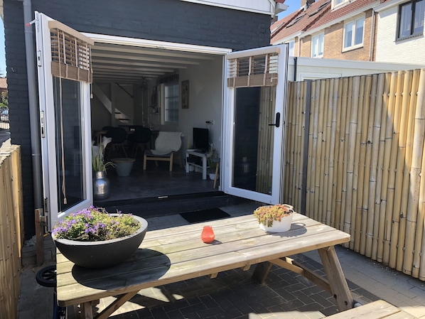 An open sliding door leading to a covered patio area with a wooden table, two potted plants, and a small orange object on the table.