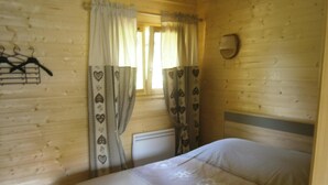 A wooden cabin room with a bed, curtains with heart patterns, a coat rack, and a wall-mounted heater.
