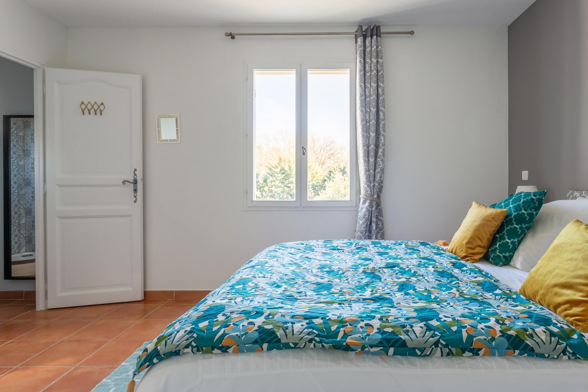 Une chambre à coucher avec un lit, une fenêtre avec des rideaux, une porte et un petit miroir accroché au mur.