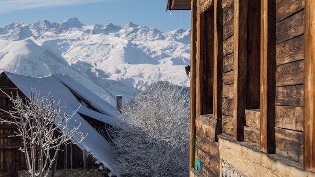 Un edificio de madera con base de piedra, un techo cubierto de nieve y una vista de montañas nevadas al fondo.