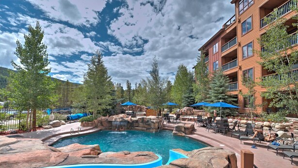 A pool area with a waterfall feature, surrounded by lounge chairs and umbrellas, and adjacent to a multi-story building. - The Springs, Extra Large Condo.  Cool winter deals! (Keystone)