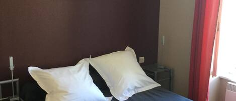 Una cama bien hecha con almohadas blancas y una colcha azul en una habitación con pared oscura, un cuadro enmarcado y una cortina roja.